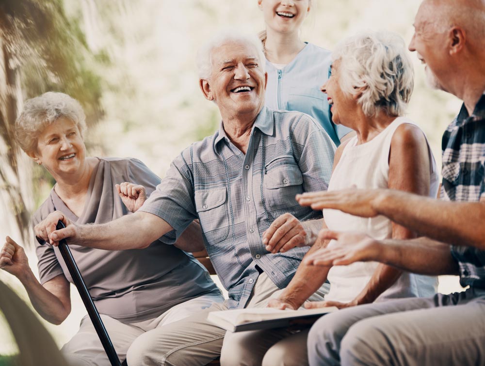 A group of four elderly residents sit outside in the sunshine and enjoy a laugh together.