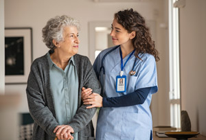An elderly woman talks a walk with a nursing assistant supporting her.