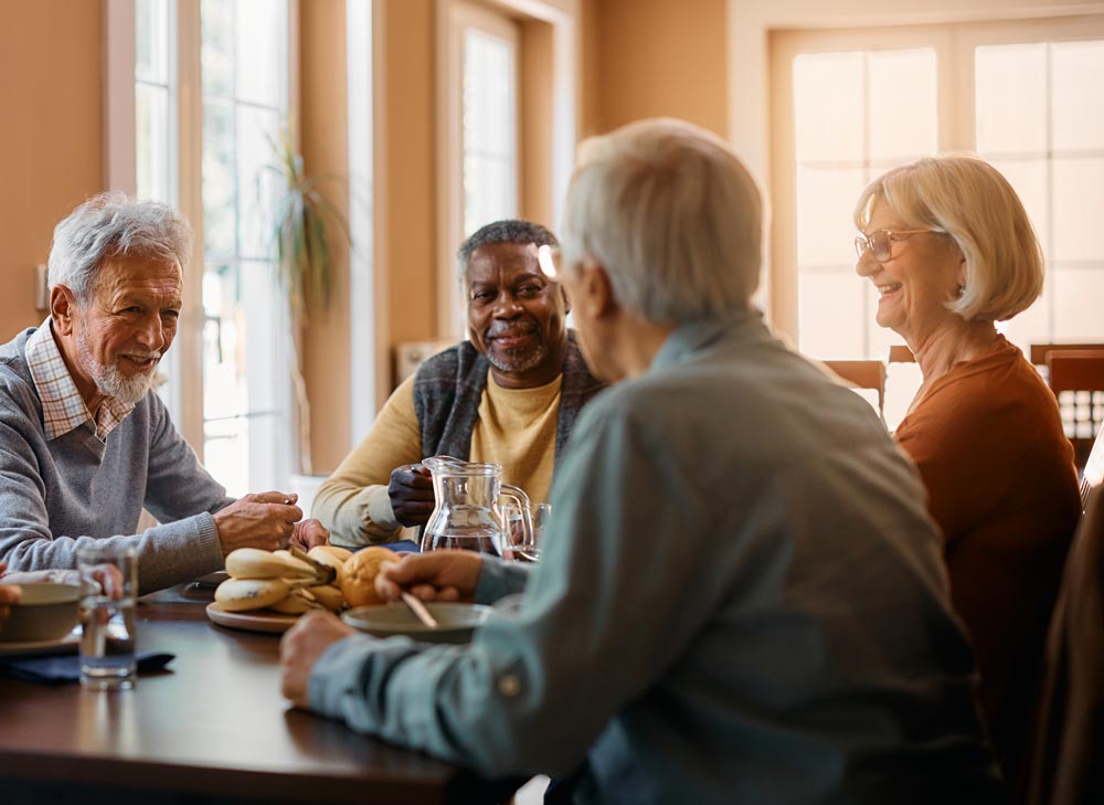A group of seniors, three men and one woman, enjoy conversation around a dining table in a well-lit resident dining hall.