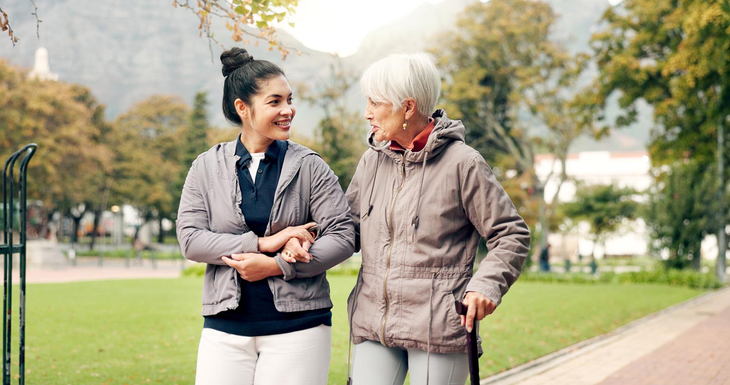 A younger woman holds an elderly woman's hand as they take a walk outside on an autumn day.